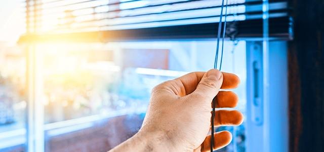 A hand adjusts window blinds with sunlight visible through window.